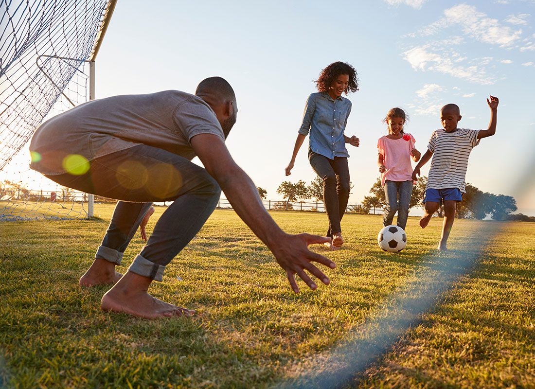 Insurance Solutions - A Boy Kicks a Football During a Game With His Family Outdoors in a Park at Sunset
